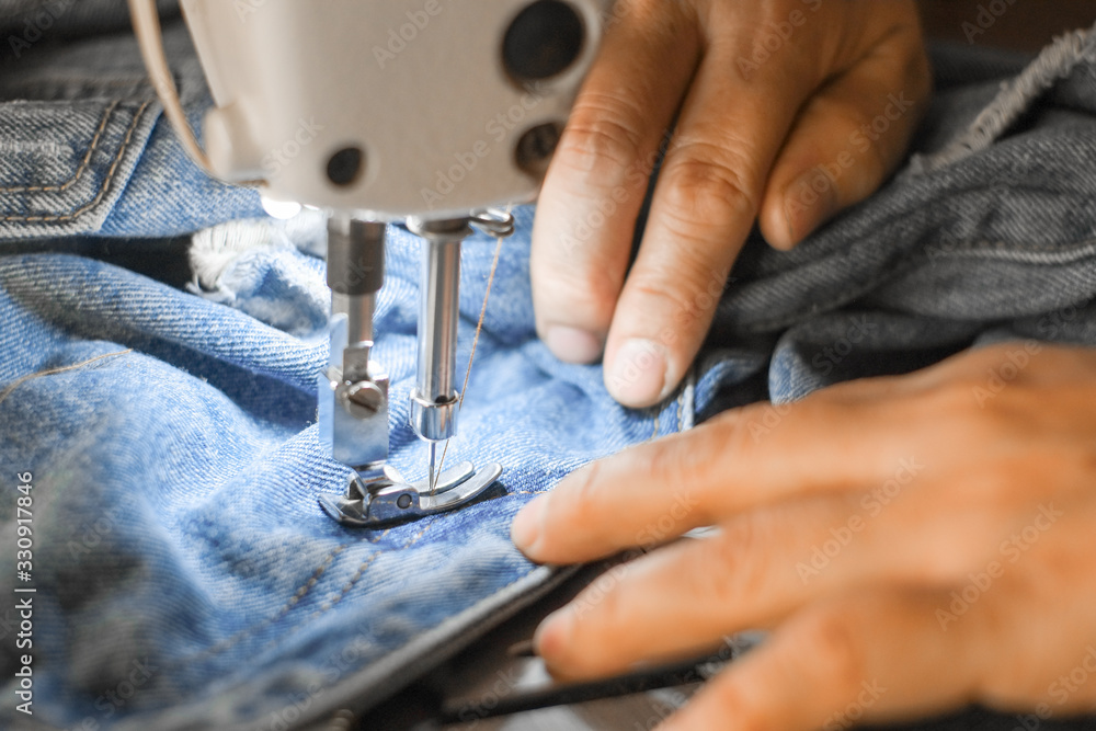 Hand of the seamstress is using a white industrial sewing machine to sew the seams of blue jeans.