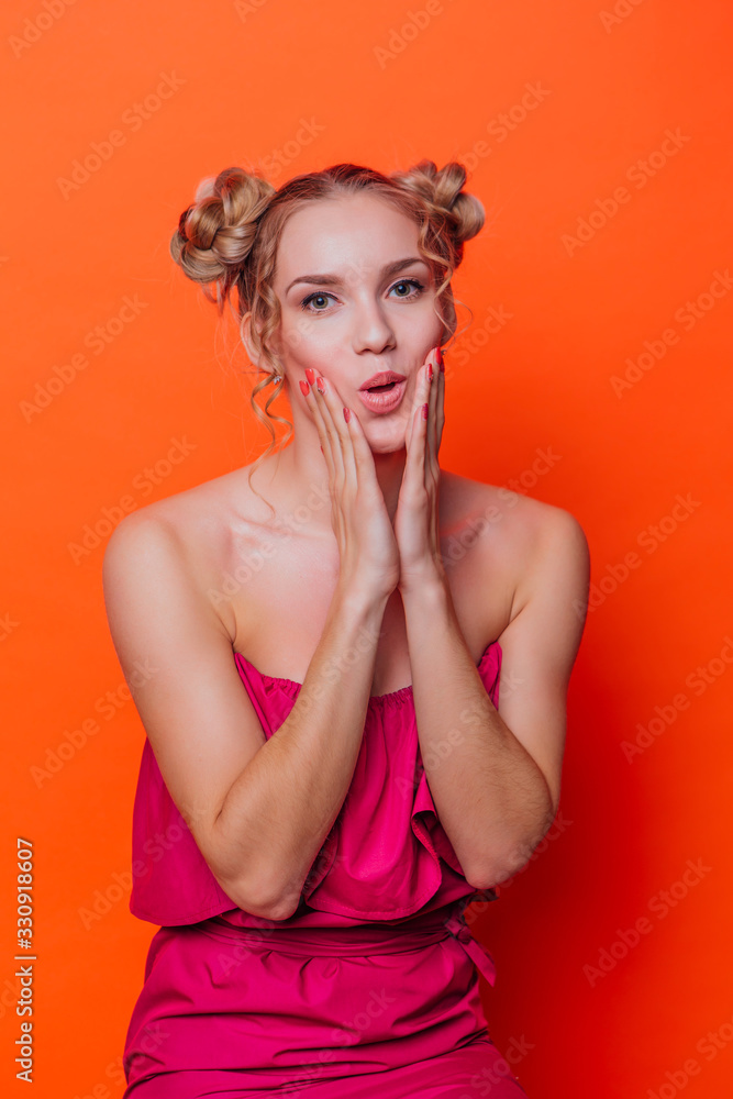 The girl in a red dress on a orange background in the studio. Blonde girl with two hair knots holding her chicks with hands The girl in a red dress on a orange background in the studio. Blonde girl with two hair knots holding her chicks with hands