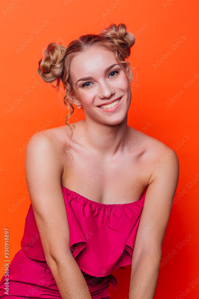 The girl in a red dress on a orange background in the studio. Blonde girl with two hair knots looking to the camera and smiling. The girl in a red dress on a orange background in the studio. Blonde girl with two hair knots looking to the camera and smiling.