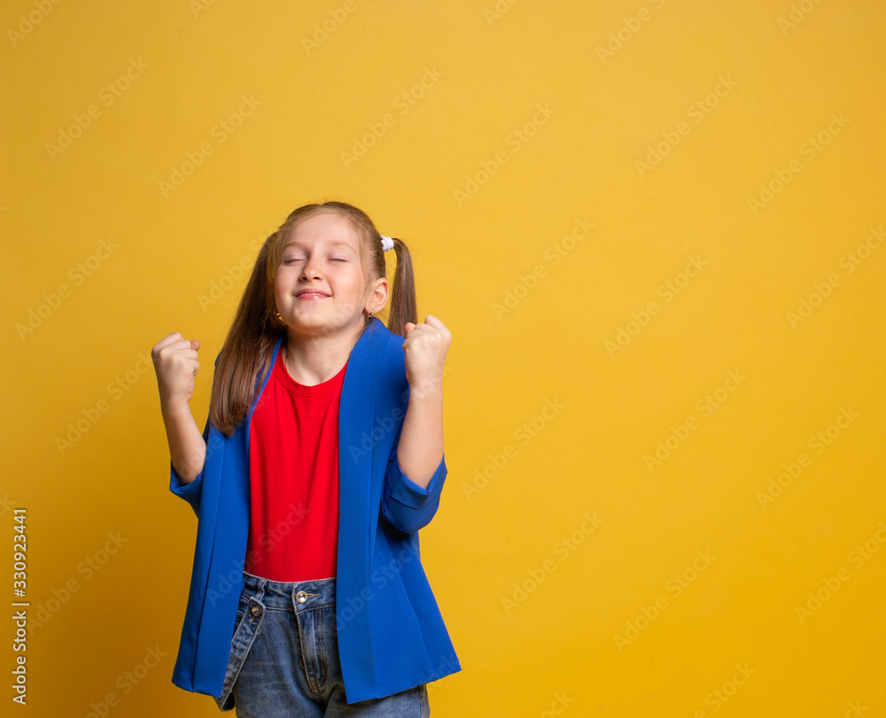 Happy joyful smiling cute little girl making a win gesture with close eyes on yellow background Happy joyful smiling cute little girl making a win gesture with close eyes on yellow background
