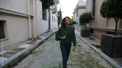 Slow motion, young woman walks at an alley looking around, bag on shoulder in Plaka, Athens, Greece, gimbal track back photo