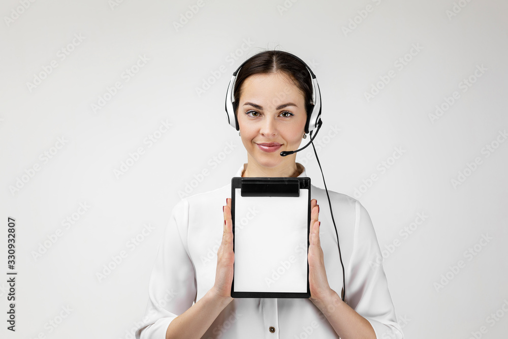 Beautiful smiling woman consultant of call center in headphones holding clipboard with blank white paper sheet on gray background. female customer support operator with headset Beautiful smiling woman consultant of call center in headphones holding clipboard with blank white paper sheet on gray background. female customer support operator with headset
