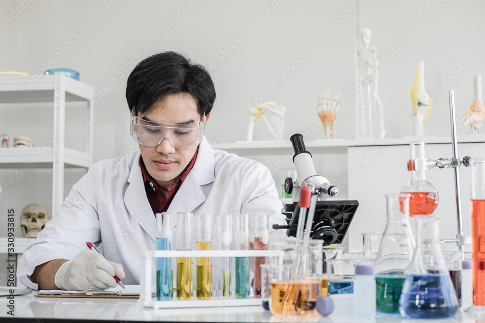 A male scientist with black hair wearing white coat and protective glassware sitting looking at test tube and solutions and a microscope in white laboratory room. A male scientist with black hair wearing white coat and protective glassware sitting looking at test tube and solutions and a microscope in white laboratory room.