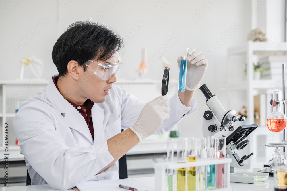 Fototapeta premium A male scientist with black hair wearing white coat and protective glassware holding magnify glass and looking at a test tube in a laboratory setting with a microscope.