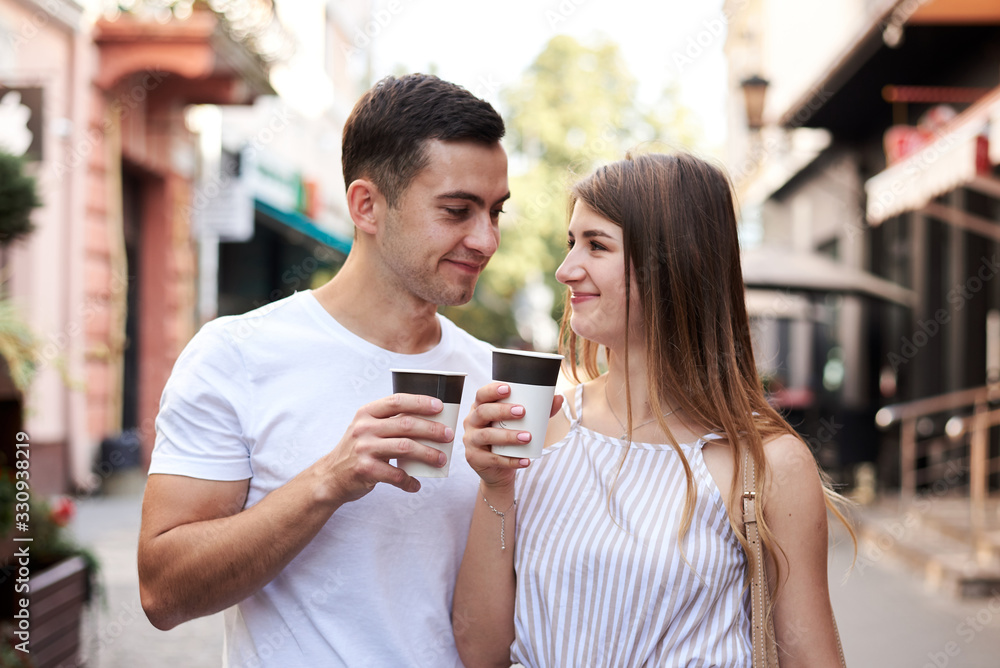 Romantic couple on date drinking coffee cappuccino in paper cups on street . Young blond woman, walking with brunette man with yummy coffee drink in summer in morning. Nutritious breakfast, snack. Romantic couple on date drinking coffee cappuccino in paper cups on street . Young blond woman, walking with brunette man with yummy coffee drink in summer in morning. Nutritious breakfast, snack.