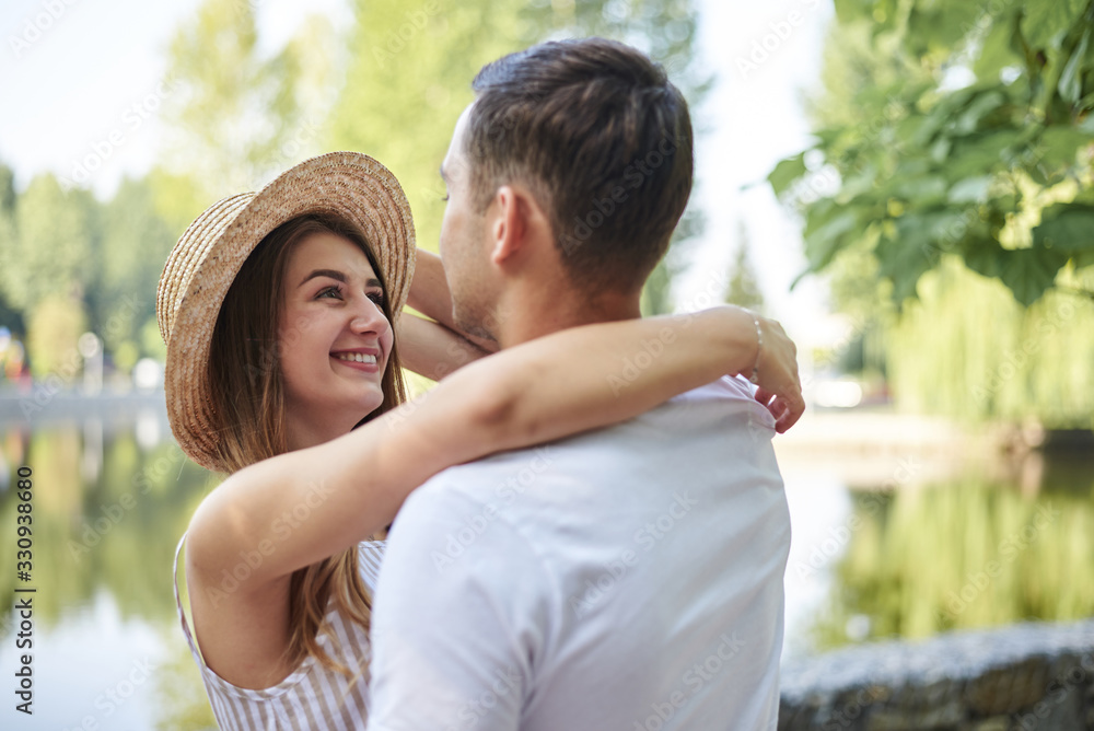 Fototapeta premium Young couple in love, hugging in the park in summer, smiling. Pretty blond girl in stripy overall and straw hat on romantic date with handsome brunette guy in white t-shirt. Sunday city walk.