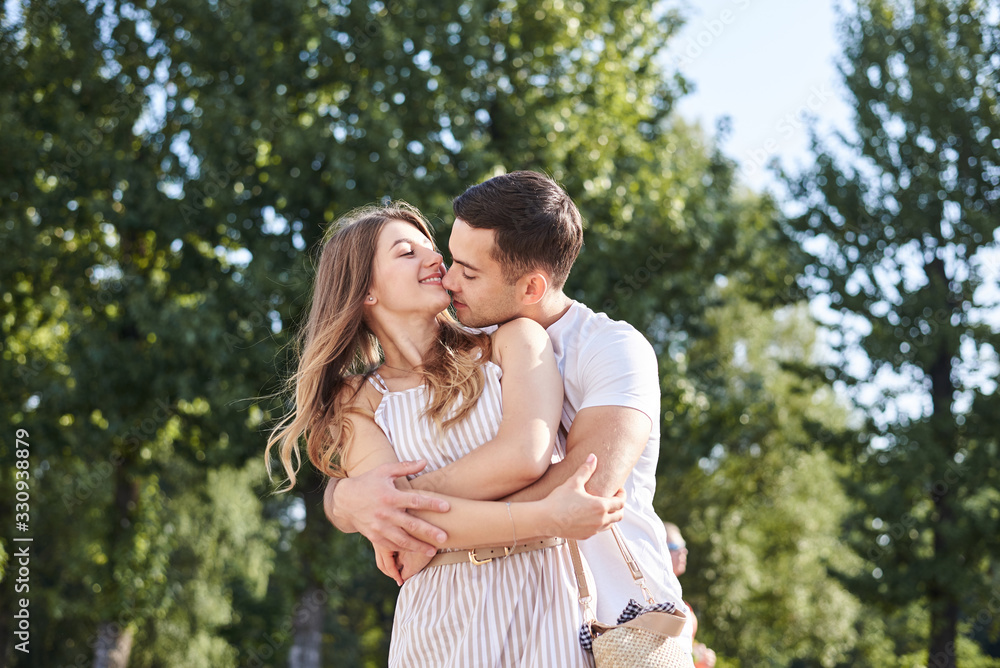 Handsome brunette guy, wearing white t-shirt, hugging and kissing pretty blond woman in stripy overall. Young couple in love, embracing in green park in summer. Romantic relationship. Valentines day. Handsome brunette guy, wearing white t-shirt, hugging and kissing pretty blond woman in stripy overall. Young couple in love, embracing in green park in summer. Romantic relationship. Valentines day.