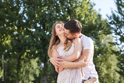 Handsome brunette guy, wearing white t-shirt, hugging and kissing pretty blond woman in stripy overall. Young couple in love, embracing in green park in summer. Romantic relationship. Valentines day.