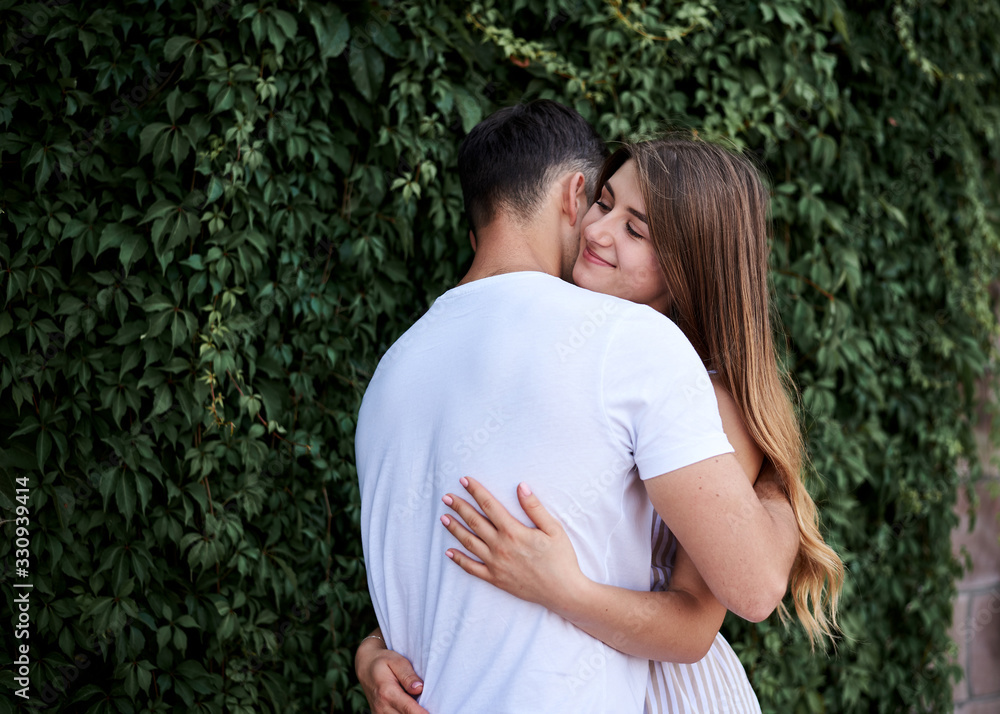 Young couple in love hugging near green bushes trees wall. Pretty blond woman, wearing stripy short overall and brunette man in white t-shirt and blue shorts on romantic date. Romantic relationship Young couple in love hugging near green bushes trees wall. Pretty blond woman, wearing stripy short overall and brunette man in white t-shirt and blue shorts on romantic date. Romantic relationship
