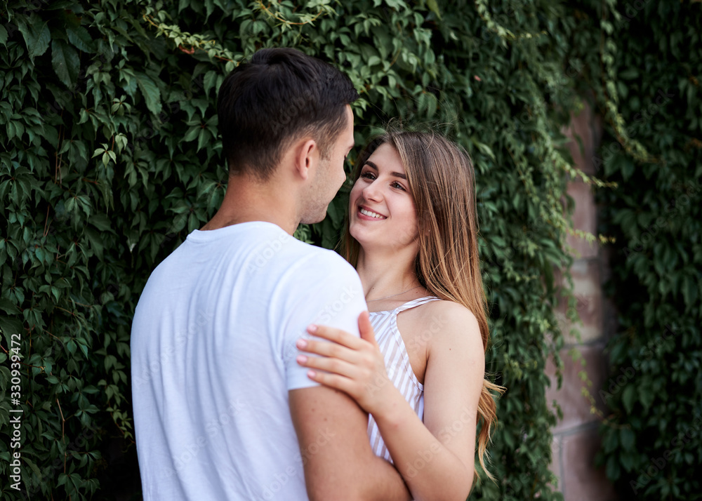 Young couple in love hugging near green bushes trees wall. Pretty blond woman, wearing stripy short overall and brunette man in white t-shirt and blue shorts on romantic date. Romantic relationship Young couple in love hugging near green bushes trees wall. Pretty blond woman, wearing stripy short overall and brunette man in white t-shirt and blue shorts on romantic date. Romantic relationship