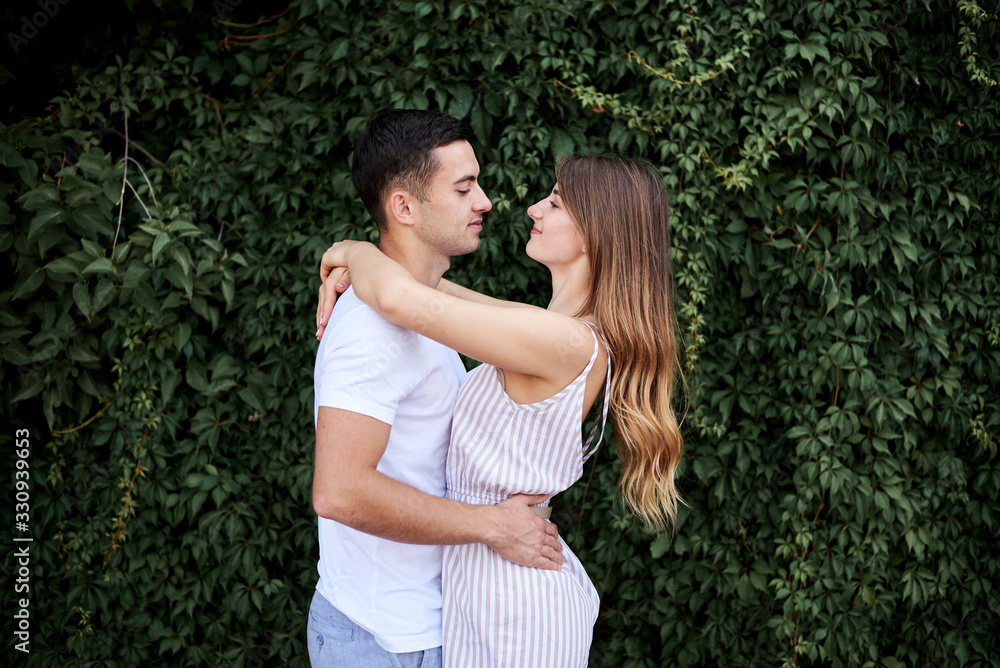 Young couple in love hugging near green bushes trees wall. Pretty blond woman, wearing stripy short overall and brunette man in white t-shirt and blue shorts on romantic date. Romantic relationship Young couple in love hugging near green bushes trees wall. Pretty blond woman, wearing stripy short overall and brunette man in white t-shirt and blue shorts on romantic date. Romantic relationship