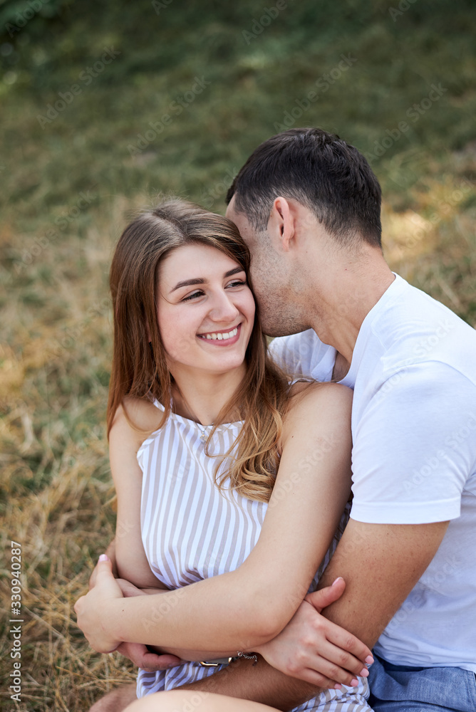 Young couple in love sitting on green yellow grass lawn hugging embracing kissing. Blond woman wearing stripy short overall and brunette man in white t-shirt blue shorts on romantic date. Relationship