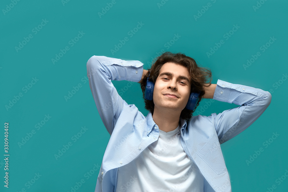 Naklejka premium Portrait of a young beautiful man wearing white t-shirt and blue shirt in blue headphones resting put his hands behind his head over blue background