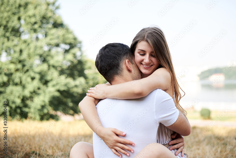 Young couple in love sitting on green yellow grass lawn hugging embracing kissing. Blond woman wearing stripy short overall and brunette man in white t-shirt blue shorts on romantic date. Relationship Young couple in love sitting on green yellow grass lawn hugging embracing kissing. Blond woman wearing stripy short overall and brunette man in white t-shirt blue shorts on romantic date. Relationship
