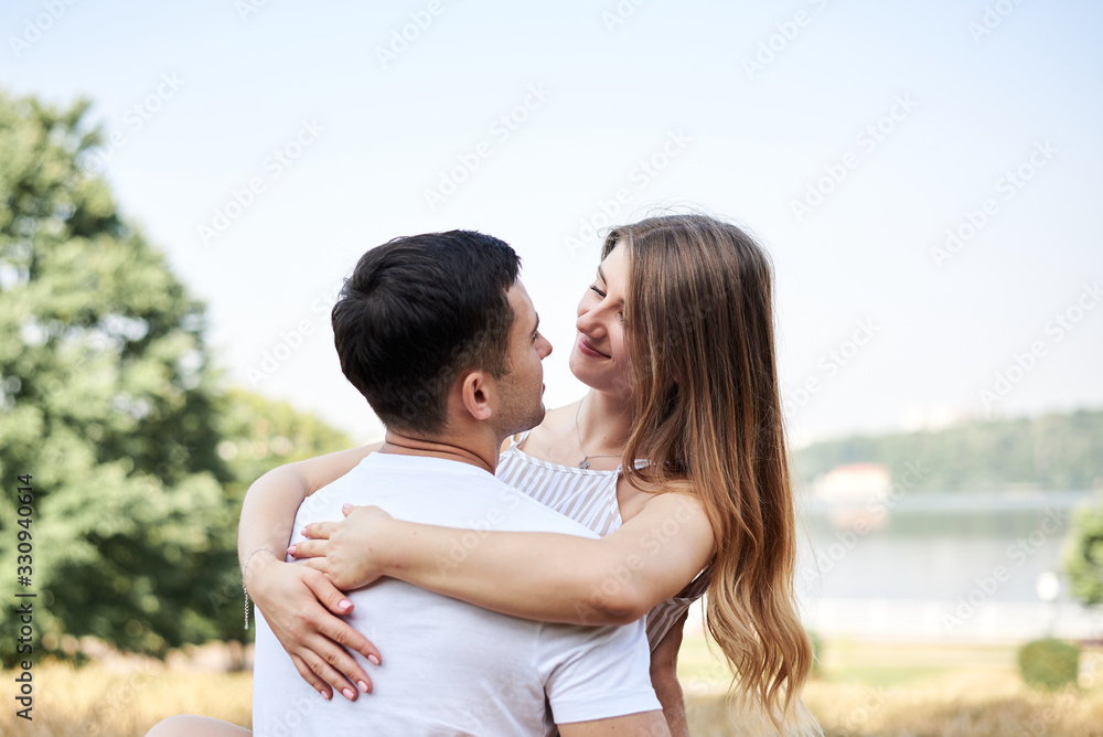 Young couple in love sitting on green yellow grass lawn hugging embracing kissing. Blond woman wearing stripy short overall and brunette man in white t-shirt blue shorts on romantic date. Relationship Young couple in love sitting on green yellow grass lawn hugging embracing kissing. Blond woman wearing stripy short overall and brunette man in white t-shirt blue shorts on romantic date. Relationship