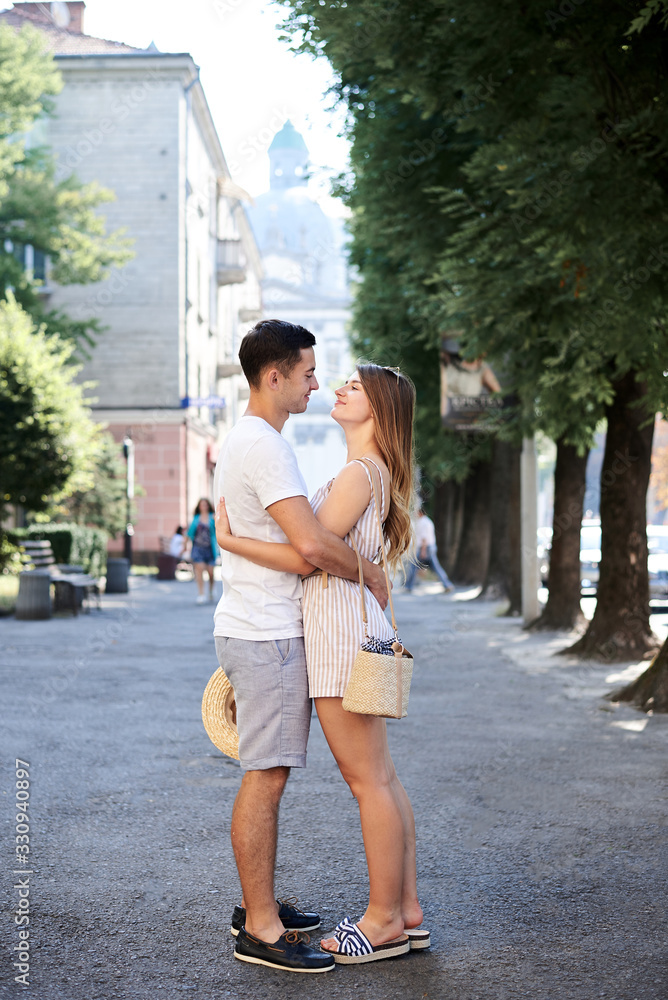 Young couple in love walking on small alley holding hands. Pretty blond woman, wearing stripy short overall and brunette man in white t-shirt and blue shorts on romantic date. Relationship. Vacation.