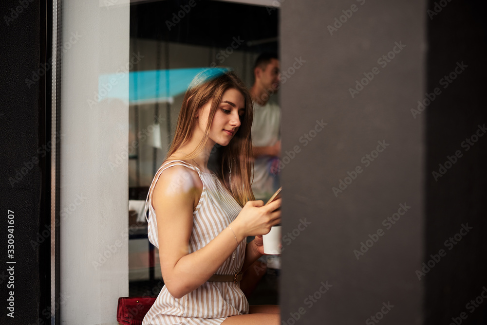 Young blond woman, wearing white overall, sitting by huge glass window in coffee shop. Pretty girl on dinner break, drinking coffee tea cappuccino in street city cafe in summer.