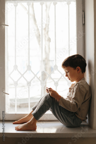 Child in home quarantine looking out of the window with his smartphone, protection against viruses during coronavirus and flu outbreak. Children and illness COVID-2019 disease concept