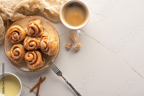 top view of fresh homemade cinnamon rolls on marble surface with cup of coffee, condensed milk, fork and cloth