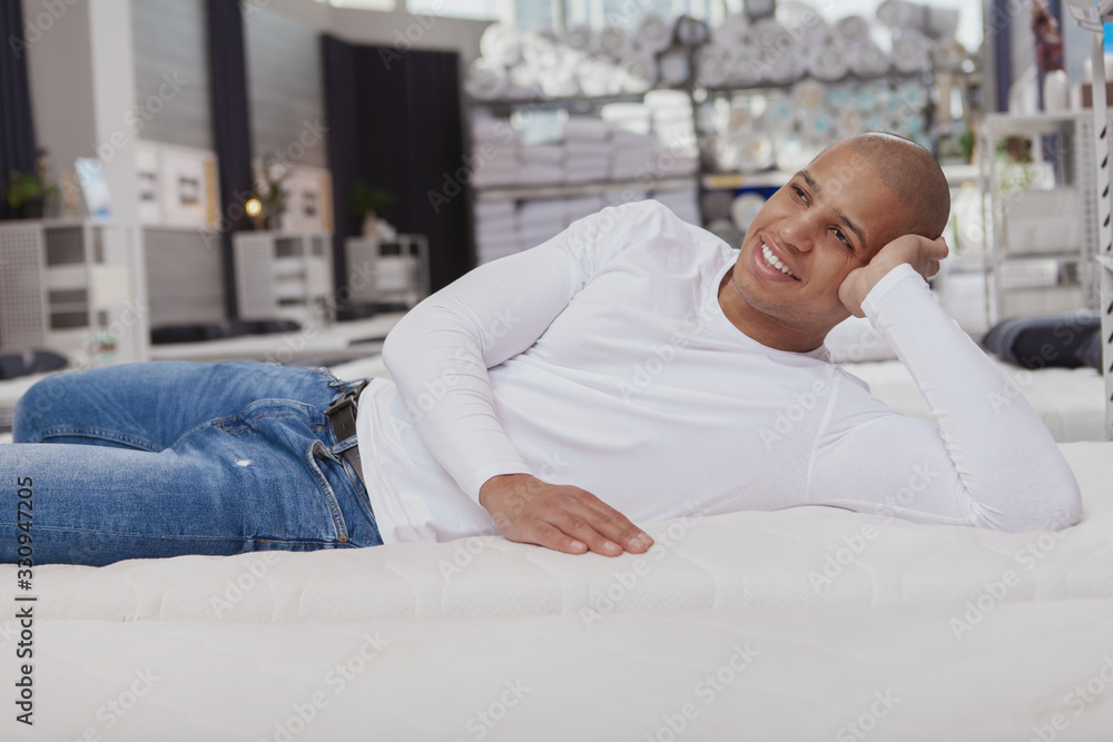Attractive young African man lying on a new bed at furniture store, smiling cheerfully. Healthy sleep concept Attractive young African man lying on a new bed at furniture store, smiling cheerfully. Healthy sleep concept