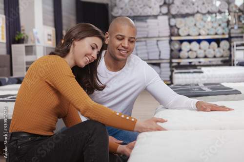 Happy young multiethnic couple choosing a new bed to buy at furniture store. Love, family, living together concept
