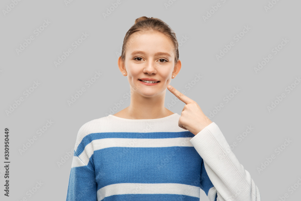 people concept - happy smiling teenage girl in pullover pointing finger to her face or showing dimple over grey background people concept - happy smiling teenage girl in pullover pointing finger to her face or showing dimple over grey background