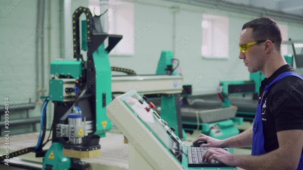 A young male operator of a CNC machine stands at the control panel in the workshop. Slider shot. A young male operator of a CNC machine stands at the control panel in the workshop. Slider shot.