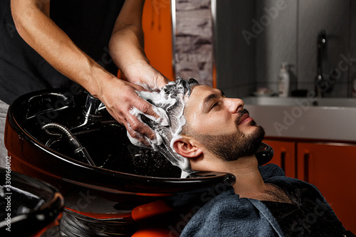 a young dark-haired guy of Indian or Asian appearance in a Barber shop on a black chair. the Barber washes his head with shampoo.