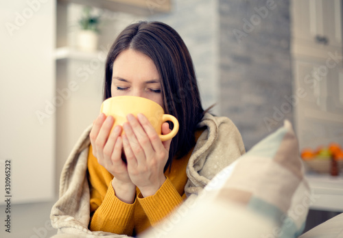 Ill young woman with flu virus at home drinking tea covered in blanket 