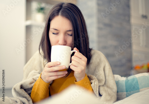 Ill young woman with flu virus at home drinking tea covered in blanket 