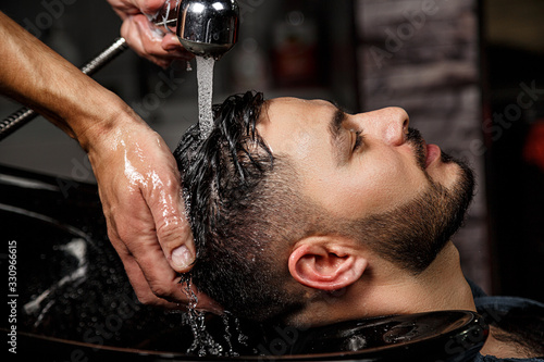 a young dark-haired guy of Indian or Asian appearance in a Barber shop on a black chair. the Barber washes his head with shampoo.