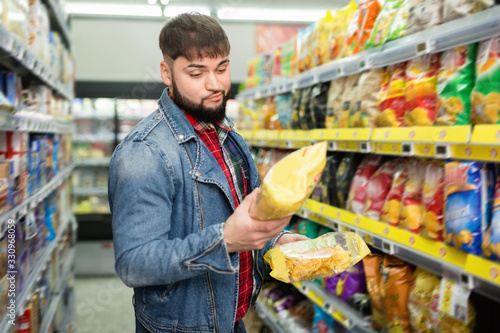 Portrait of doubting bearded guy choosing potato chips at grocery store