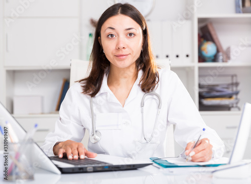 Practicioner girl in medical uniform is working with documents and laptop