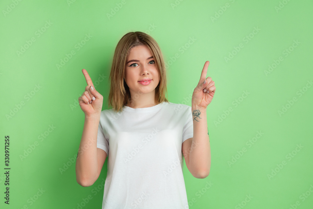 Pointing up. Caucasian young woman's portrait isolated on green studio background. Beautiful female model in white shirt. Concept of human emotions, facial expression, sales, ad, youth. Pointing up. Caucasian young woman's portrait isolated on green studio background. Beautiful female model in white shirt. Concept of human emotions, facial expression, sales, ad, youth.