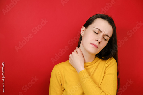Tired female feeling stiff sore neck pain concept rubbing massaging tensed muscles suffer from fibromyalgia, has gloomy expression, wears sweater, isolated on red background. Fibromyalgia concept