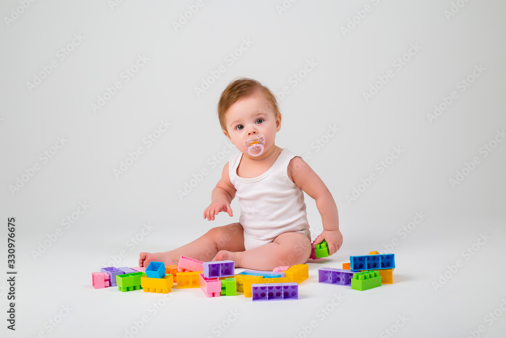 baby with a pacifier in his mouth in white clothes sitting playing with a multi-colored constructor on a white background, space for text baby with a pacifier in his mouth in white clothes sitting playing with a multi-colored constructor on a white background, space for text