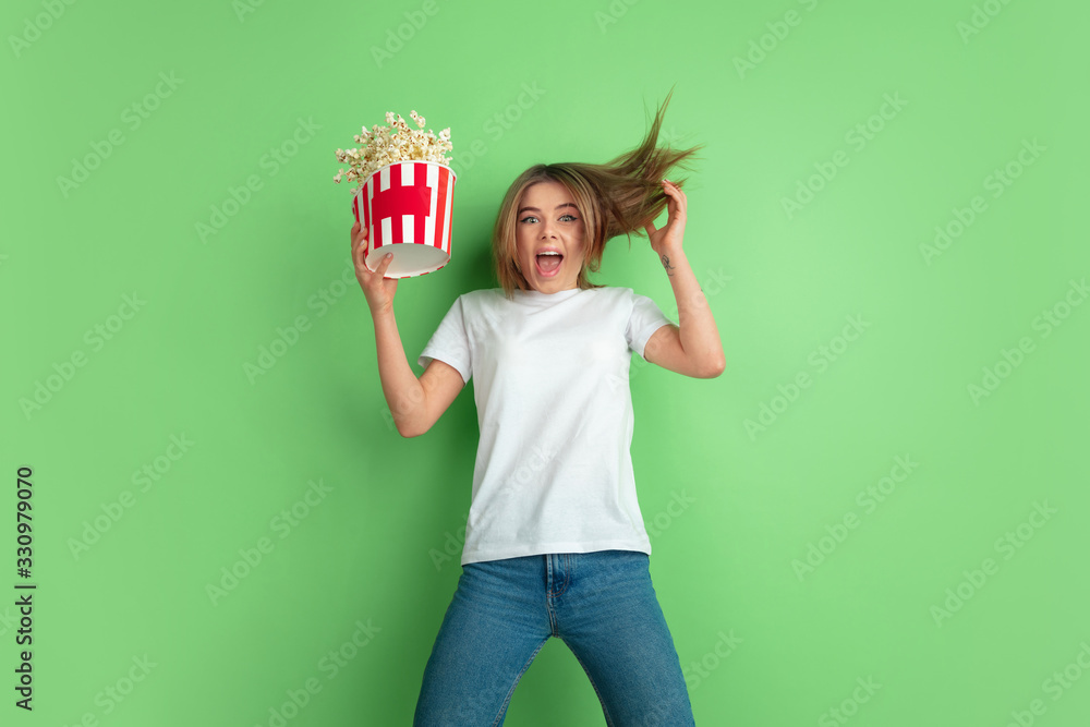 Screaming, popcorn's flying. Caucasian young woman's portrait isolated on green studio background. Beautiful female model in white shirt. Concept of human emotions, facial expression, sales, ad, youth