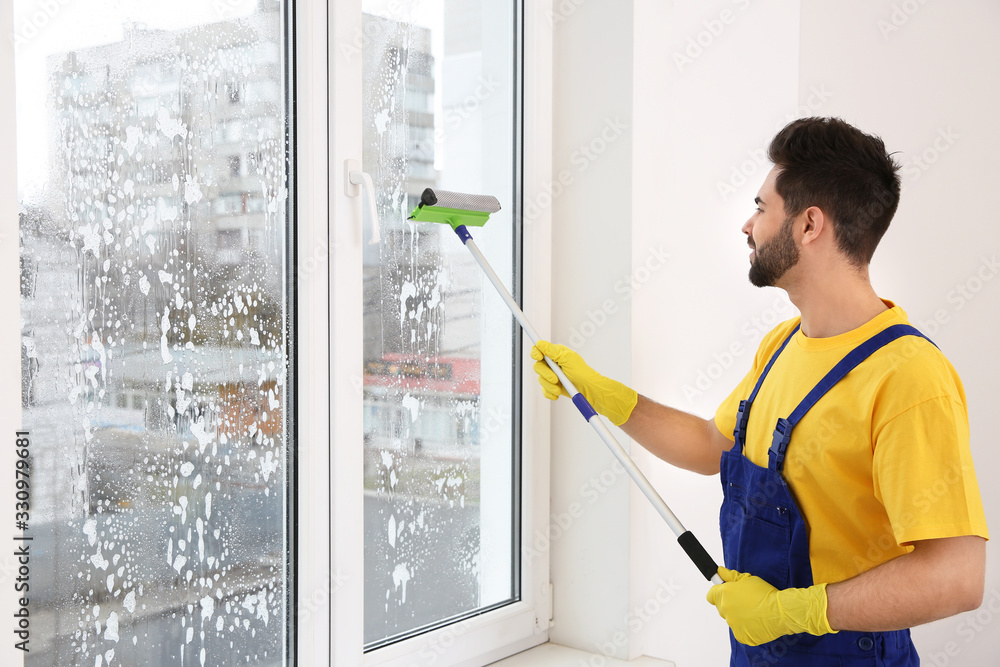 Professional young janitor in uniform cleaning window indoors Professional young janitor in uniform cleaning window indoors
