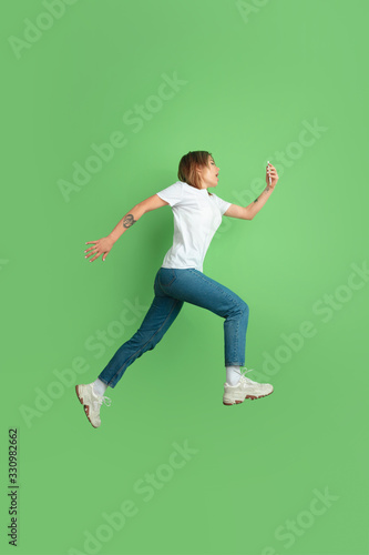 Shocked using phone in jump. Caucasian young woman's portrait isolated on green studio background. Beautiful female model in white shirt. Concept of human emotions, facial expression, sales, ad, youth