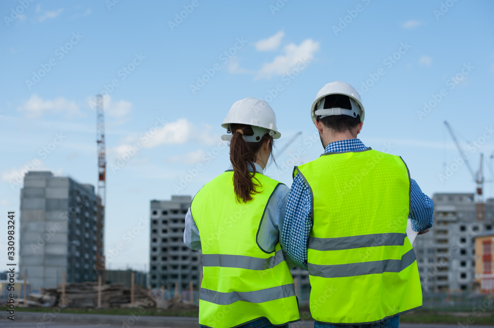 Rear View - Two Young Engineers A Man And A Woman In Green Vests And Helmets Are Discussing A Plan Rear View - Two Young Engineers A Man And A Woman In Green Vests And Helmets Are Discussing A Plan