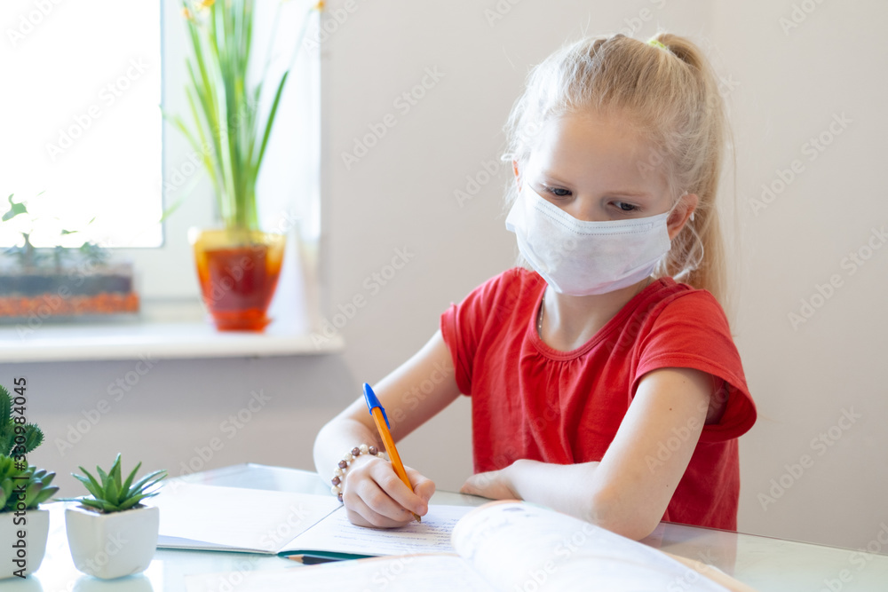 Serios sad blonde schoolgirl in medical mask stydying at home, doing school homework, writing in notebook. Reading training books on table. Back to school. Distance learning online education