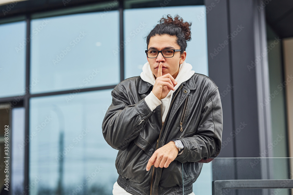 Handsome young man with curly black hair and in glasses is on the street against building Handsome young man with curly black hair and in glasses is on the street against building
