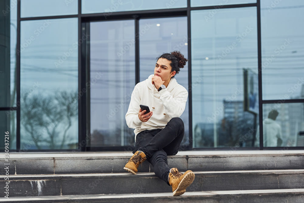 Handsome young man with curly black hair and with smartphone in hands is on the street sitting on the stairs against building Handsome young man with curly black hair and with smartphone in hands is on the street sitting on the stairs against building