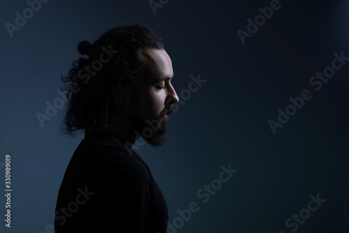 studio dramatic profile portrait of a young bearded guy of thirty years old, with long hair gathered, looks down. On a dark background. A ray of light on the face. Dramatic light