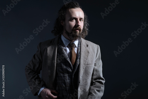 studio dramatic of a young bearded and curly-haired guy of thirty years, in an expensive business suit, holds his hand by the lapel of his jacket. On a dark background.