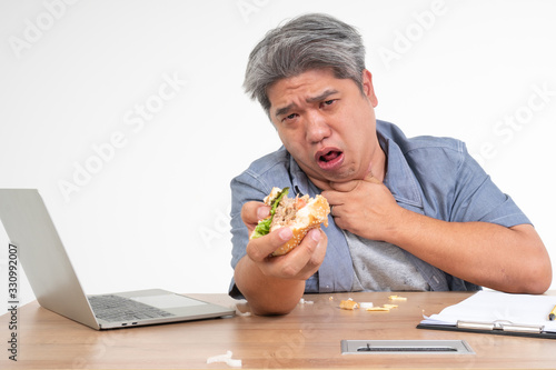 Bild auf Leinwand Asian man working and eating a burger on office desk and holding his neck after choking foods