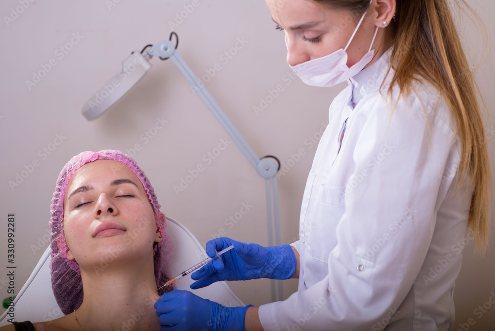 Naklejka premium portrait in the cosmetologists office. A young girl in a medical suit, gloves and a syringe performs an injection into the facial and neck muscles to treat wrinkles. scars, muscle spasm, torticollis