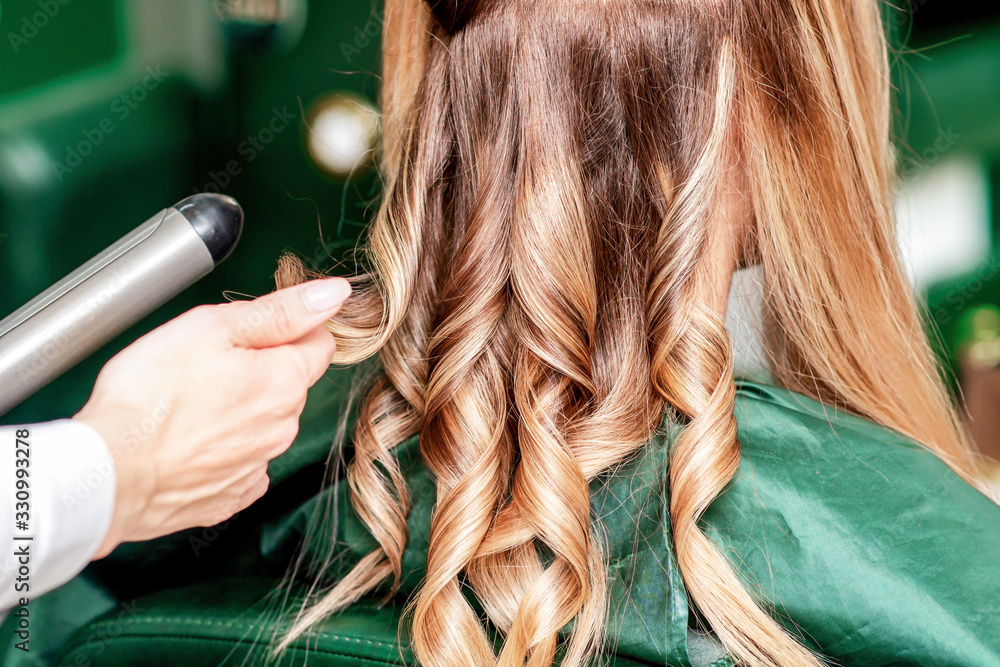 Fototapeta premium Hairdresser twists hair of woman by curling iron close up.