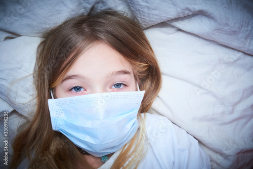 Sick girl lying in bed with protection mask on face against infection. Selective focus