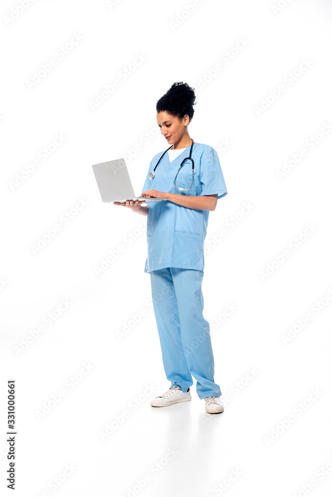 Full length view of african american nurse with stethoscope using laptop on white background Full length view of african american nurse with stethoscope using laptop on white background